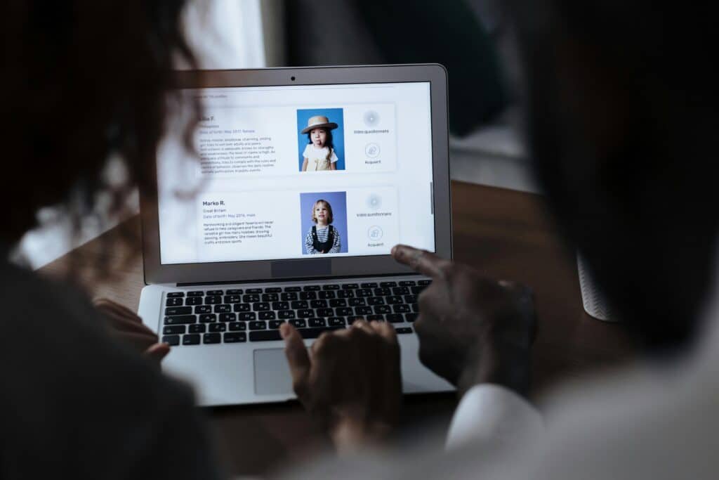 Two adults reviewing child adoption profiles on a laptop screen, considering potential matches.
